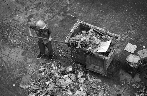 A worker wearing a protective helmet and casual clothing is standing on a paved outdoor surface next to a large open-top industrial waste skip, which is filled with mixed refuse including cardboard, plastic bags, and various discarded items. The worker is holding a long-handled tool, possibly a rake, and is in the process of sorting or clearing debris around the skip. The area around the skip shows scattered trash and debris, with some waste materials spilled onto the ground, indicating a rubbish removal task. In the background, a small metal trolley and additional waste materials are visible, suggesting an active waste collection or disposal environment consistent with commercial waste management services, such as those offered by Commercial Waste Woking. The scene is in a neutral outdoor setting, likely a yard or driveway associated with waste management activity, with natural or ambient lighting illuminating the area.