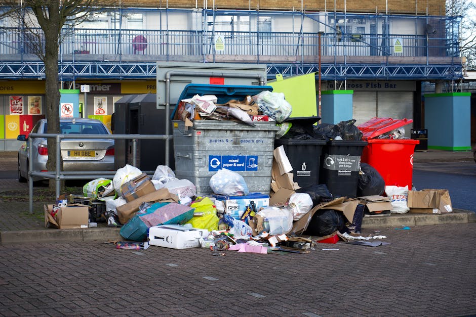 A large collection of mixed waste and rubbish overflowing from a communal bin situated on a paved street in a commercial or residential area. The bin is a standard grey, with a blue recycling symbol, 