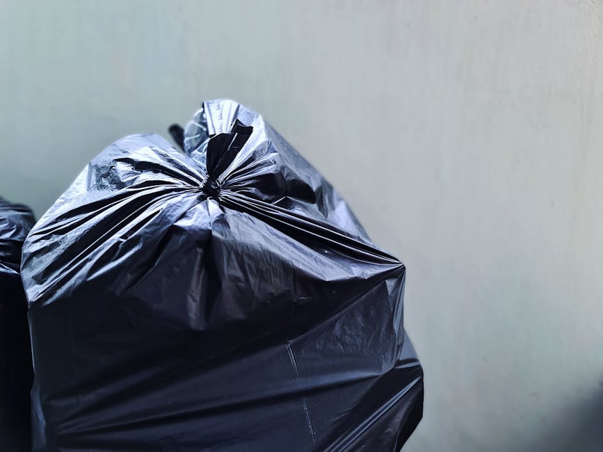 A large collection of mixed waste and rubbish overflowing from a communal bin situated on a paved street in a commercial or residential area. The bin is a standard grey, with a blue recycling symbol, 