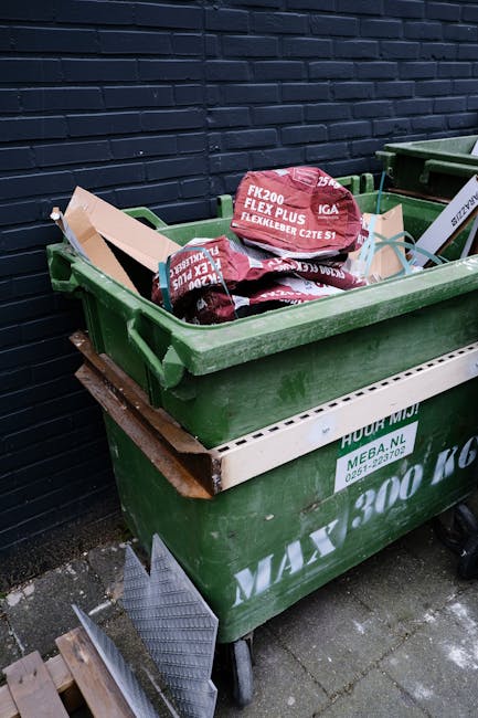 A large green plastic waste container filled with red and black bags labeled 'FK200 FLEXY PLUS', situated outdoors on a paved surface against a dark-colored brick wall. The container is placed on a wh