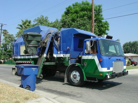 A commercial rubbish collection truck is parked on a residential street with a clear blue sky overhead. The truck has a large, rectangular, blue metal body for waste collection, with a green stripe along the lower section and a silver front cab with large side mirrors and a prominent windshield. The rear of the truck features an automated lifting mechanism for bins, with a green waste bin positioned on the left side, partially lifted to load waste. The bin is made of durable plastic with a textured surface, and appears to be filled with general waste. The street is lined with lush green trees and grass, with a few residential houses visible in the background, indicating a typical suburban area. The scene is well-lit by natural daylight, emphasizing the clean, reflective surfaces of the truck and the vibrant colors of the environment. This image exemplifies waste collection services, such as those provided by Commercial Waste Woking, operating efficiently within the local area, including Woking and surrounding postcode areas.