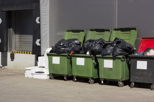 A row of four large outdoor rubbish bins placed on a concrete surface, with two green bins and one black bin visible, all filled with black rubbish bags. The green bins are positioned with their lids open, revealing bags inside, while the black bin is closed. To the left, white concrete blocks are stacked in front of a building with a roller shutter door and painted safety markings. In the background, part of a grey building facade is visible, indicating a commercial or industrial area, possibly near Woking. The scene appears to be part of a waste collection or disposal site managed by Commercial Waste Woking, reflecting typical commercial rubbish management practices in the local area.
