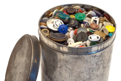 A cylindrical metal container filled with a large variety of buttons in different sizes, shapes, and colours, including black, white, blue, green, and beige, with some buttons featuring metallic finishes. The container is situated outdoors on a paved surface, with a slightly worn, weathered appearance and an open lid resting beside it. The background is plain white, emphasizing the focus on the container and its contents, which are associated with waste collection and rubbish removal services in a domestic or commercial setting nearby Woking. The detailed textures of the metal surface and the shiny, smooth surfaces of the buttons are clearly visible, supporting the visual context of rubbish disposal that commercial waste management services might handle.